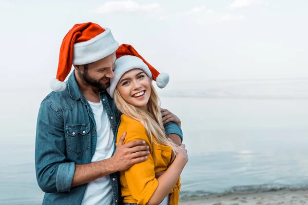 Sorrindo namorado em santa chapéu abraçando namorada na praia — Fotografia de Stock