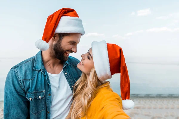 Sorridente coppia in cappelli di Babbo Natale abbracciarsi sulla spiaggia e guardarsi l'un l'altro — Foto stock