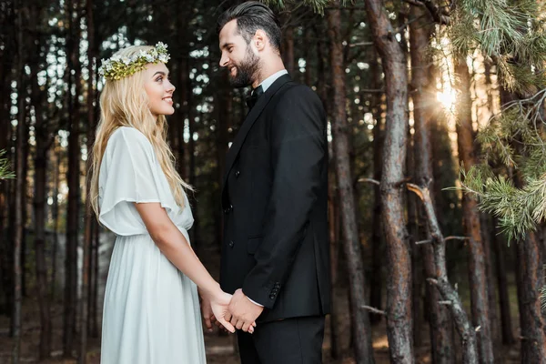 Side view of happy wedding couple holding hands and looking at each other during sunset in forest — Stock Photo