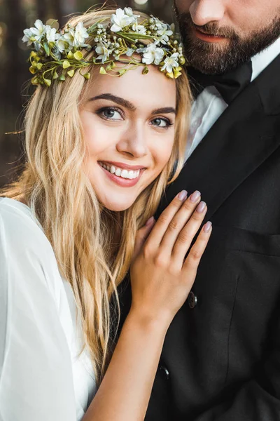 Smiling attractive bride in wreath hugging groom and looking at camera — Stock Photo