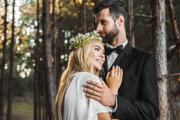 Hermosa novia sonriente en vestido blanco y novio guapo en traje abrazándose en el bosque - foto de stock