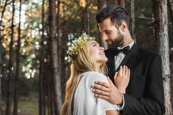 Beautiful bride in white dress and handsome groom in suit hugging in forest and looking at each other — Stock Photo
