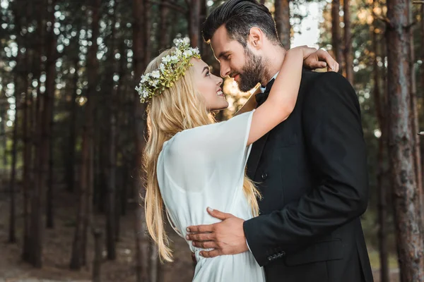 Beautiful happy bride in white dress and handsome groom in suit hugging and going to kiss in forest — Stock Photo