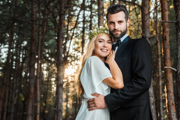 Низький кут зору усміхненої весільної пари, що обіймається і дивиться на камеру під час заходу сонця в лісі — стокове фото