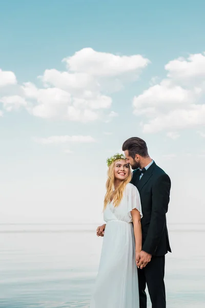 Wedding couple in suit and white dress holding hands and looking at each other on beach — Stock Photo