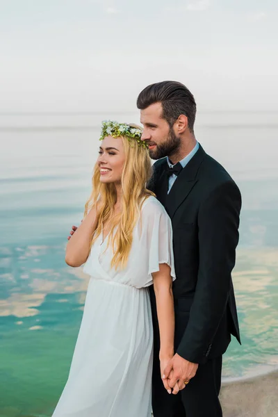 Hochzeitspaar in Anzug und weißem Kleid hält Händchen und schaut am Strand weg — Stockfoto