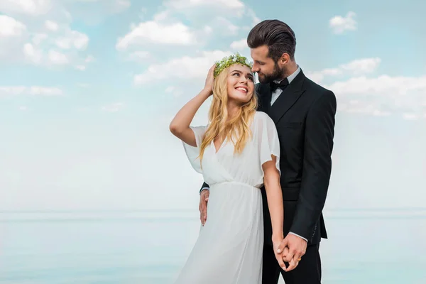 Couple de mariage affectueux en costume et robe blanche tenant la main et se regardant sur la plage — Photo de stock