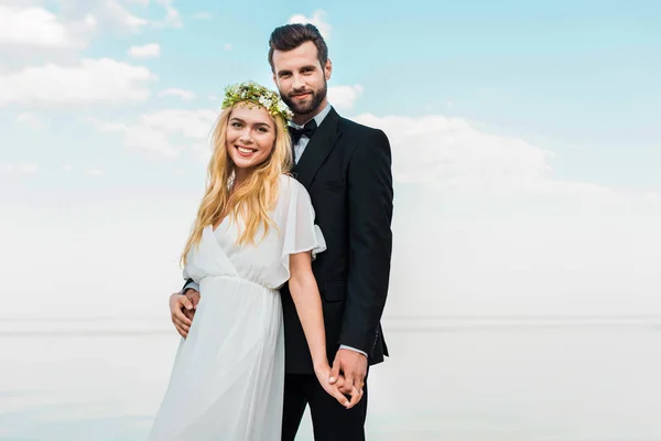 Coppia di nozze in abito bianco e abito che si tiene per mano e guardando la fotocamera sulla spiaggia — Foto stock