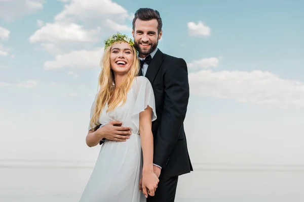 Смеющаяся свадебная пара в костюме и белом платье, держащиеся за руки и смотрящие в камеру на пляже — стоковое фото