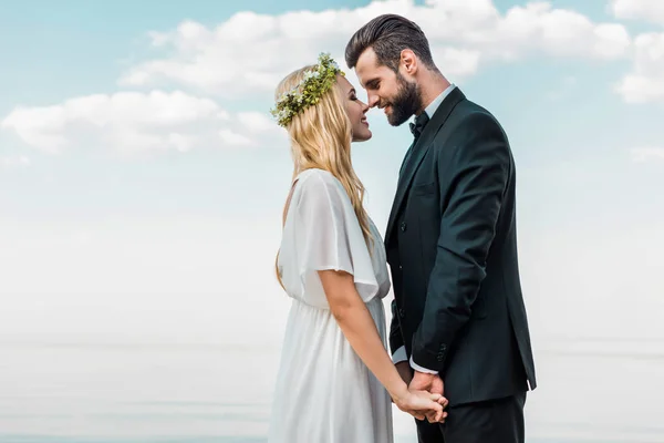 Wedding couple in suit and white dress holding hands and touching with noses on beach — Stock Photo