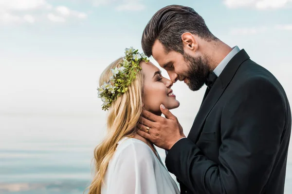 Happy wedding couple in suit and white dress touching with noses on beach — Stock Photo