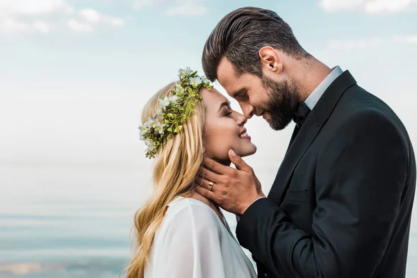 Feliz pareja de boda en traje y vestido blanco tocando con narices en la playa - foto de stock