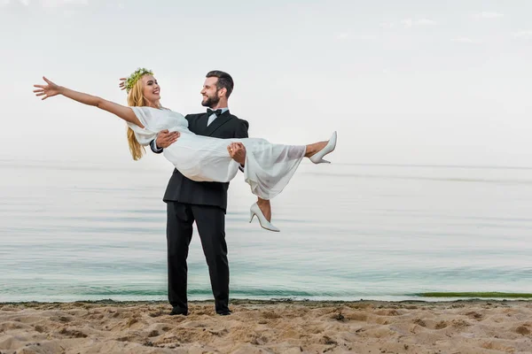 Novio guapo en traje sosteniendo atractiva novia sonriente en vestido blanco en la playa - foto de stock