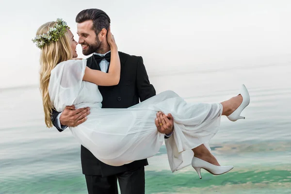 Noivo bonito em terno segurando noiva atraente em vestido branco na praia, eles olhando um para o outro — Fotografia de Stock