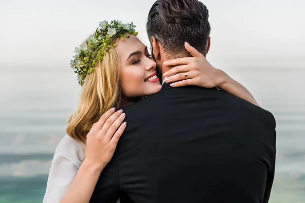 Atractiva novia con los ojos cerrados y el novio en traje abrazándose en la playa - foto de stock