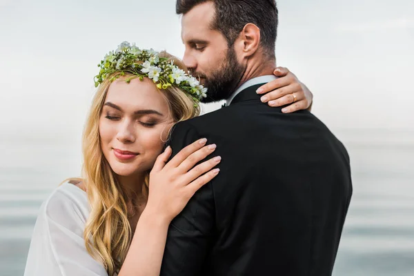 Atractiva novia con los ojos cerrados y el novio en traje abrazándose en la playa - foto de stock