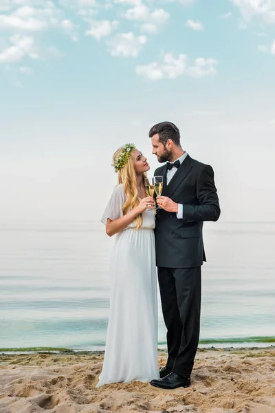 Pareja de boda tintineo con copas de champán en la playa y mirando el uno al otro - foto de stock