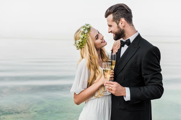 Pareja de boda sosteniendo copas de champán y mirándose en la playa - foto de stock