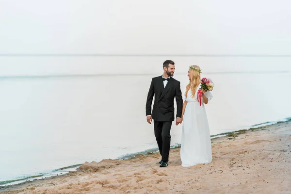 Pareja de boda tomados de la mano, caminando y mirándose en la playa - foto de stock