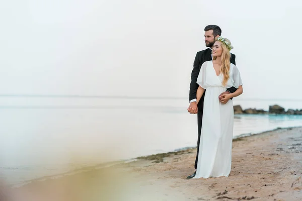 Noivo bonito em terno abraçando noiva atraente em vestido branco na praia — Fotografia de Stock