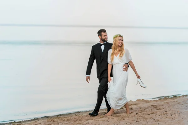 Casamento casal abraçando, andando na praia e olhando para longe, noiva segurando saltos altos na mão — Fotografia de Stock