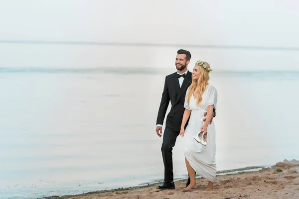 Улыбающаяся свадебная пара обнимается и гуляет по пляжу, невеста держит высокие каблуки в руке — стоковое фото