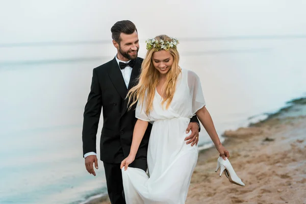 Happy wedding couple hugging and walking on beach, bride holding high heels in hand — Stock Photo