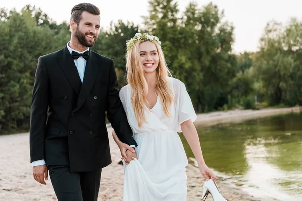 Sonriente pareja de boda tomados de la mano y caminando en la playa, novia sosteniendo tacones altos en la mano - foto de stock