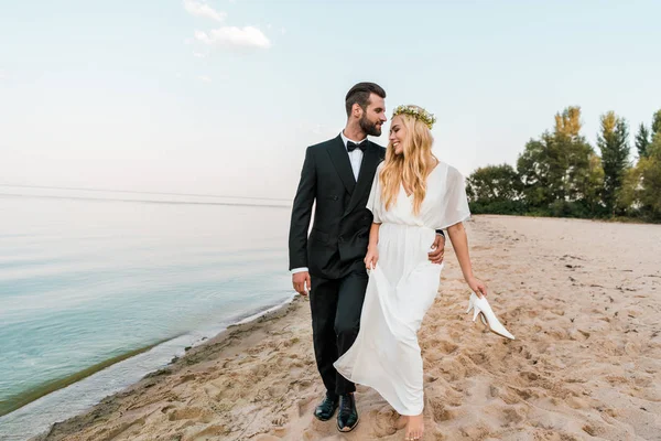 Свадебная пара обнимается и гуляет по песчаному пляжу, невеста держит высокие каблуки в руке — стоковое фото