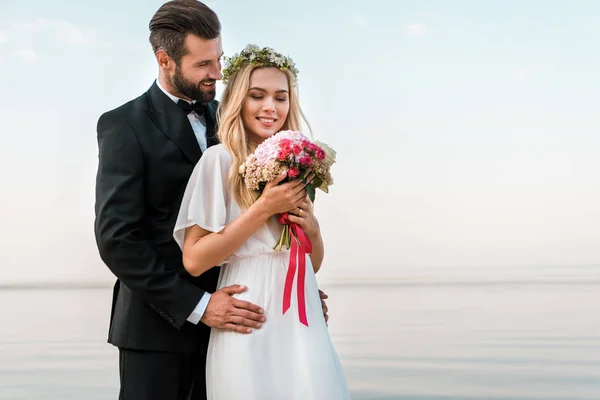 Groom hugging bride and she holding wedding bouquet on beach — Stock Photo