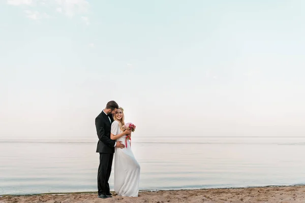 Groom hugging bride and she holding wedding bouquet on sandy ocean beach — Stock Photo