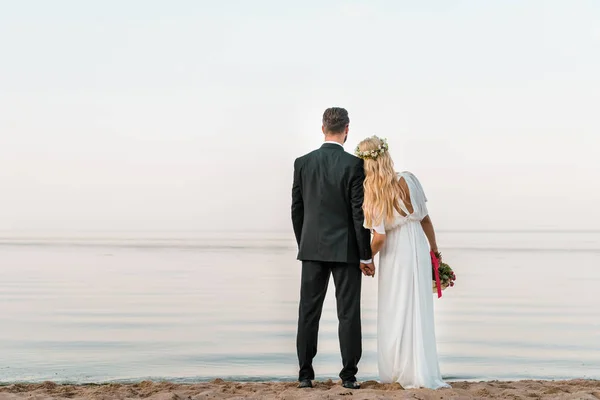 Vue arrière du couple de mariage debout sur la plage avec bouquet de mariage et regardant la mer — Photo de stock