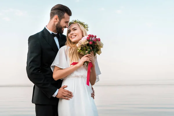 Bräutigam umarmt lächelnde Braut und sie hält Brautstrauß am Strand — Stockfoto