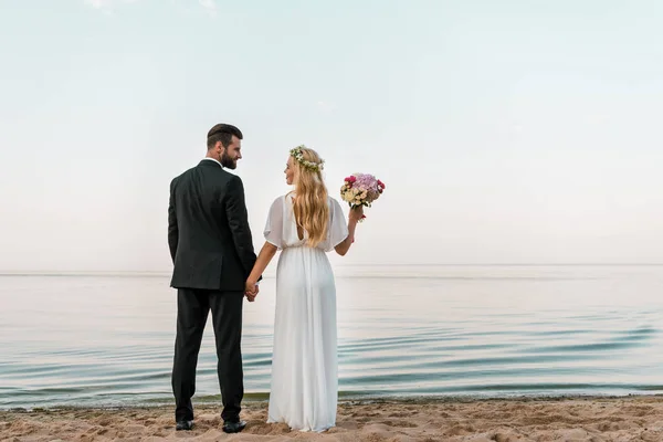 Vue arrière du couple de mariage debout sur la plage avec bouquet de mariage et se regardant — Photo de stock
