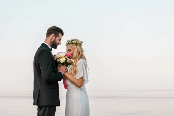 Vista lateral do casal de casamento de pé com buquê na praia, noiva farejando rosas — Fotografia de Stock