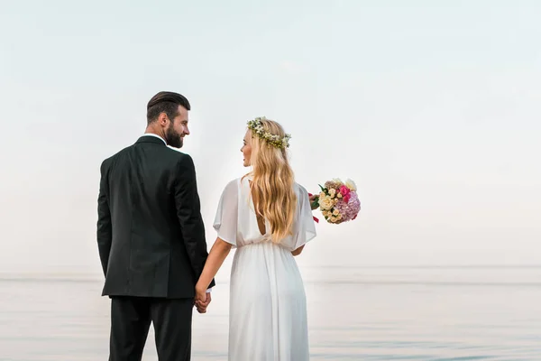Vista trasera de la pareja de boda de pie en la playa, tomados de la mano y mirando el uno al otro - foto de stock