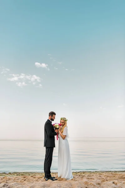 Вид сбоку свадебной пары стоящей с букетом на песчаном пляже, невеста нюхающая розы — стоковое фото