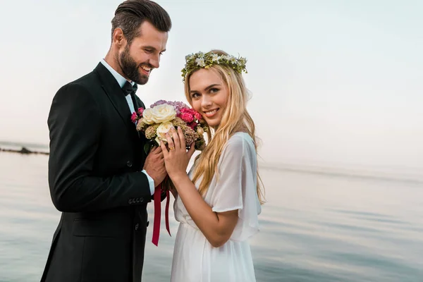 Side view of wedding couple standing with bouquet on beach, smiling bride looking at camera — Stock Photo