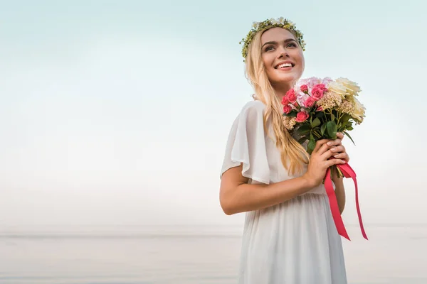 Усміхнена приваблива наречена в білій сукні і вінку, що тримає весільний букет на пляжі — стокове фото
