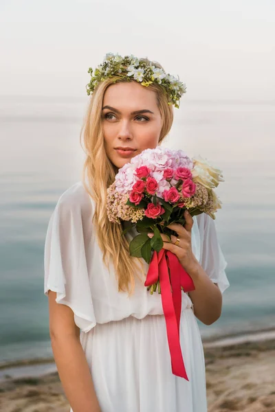 Noiva atraente em vestido branco e grinalda segurando buquê de casamento na praia de areia e olhando para longe — Fotografia de Stock