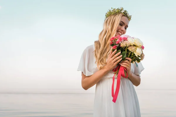 Novia atractiva sonriente en vestido blanco y corona mirando ramo de boda en la playa - foto de stock