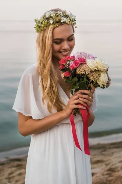 Attractive bride in white dress and wreath sniffing wedding bouquet on beach — Stock Photo
