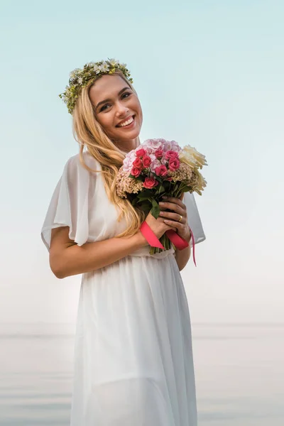 Sonriente hermosa novia en vestido blanco y corona celebración de ramo de boda en la playa y mirando a la cámara - foto de stock