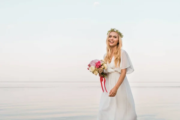 Sorridente sposa attraente in abito bianco e ghirlanda tenendo bouquet da sposa e camminando vicino al mare — Foto stock