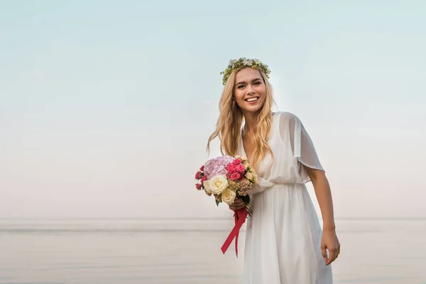 Lächelnde attraktive Braut in weißem Kleid und Kranz mit Hochzeitsstrauß aus Rosen und Blick in die Kamera am Strand — Stockfoto