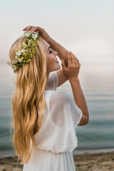 Side view of attractive bride in white dress and wreath of flowers standing with closed eyes on beach — Stock Photo