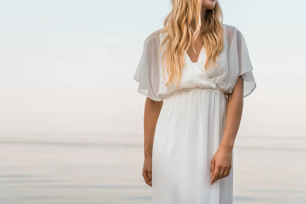 Imagen recortada de novia en vestido blanco elegante de pie cerca del océano - foto de stock