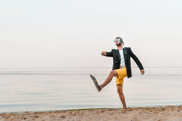 Uomo in giacca nera e pantaloncini che cammina con maschera da nuoto e pinne sulla spiaggia sabbiosa — Foto stock