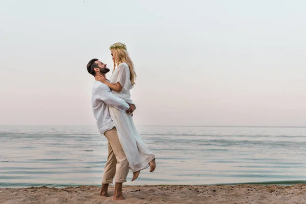 Visão lateral do noivo segurando noiva e eles olhando um para o outro na praia — Fotografia de Stock
