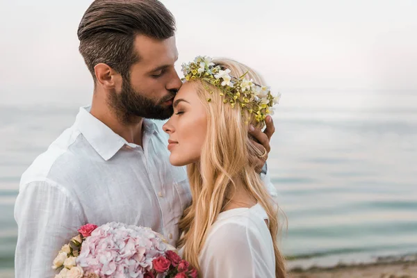 Side view of handsome groom kissing attractive bride forehead on beach — Stock Photo